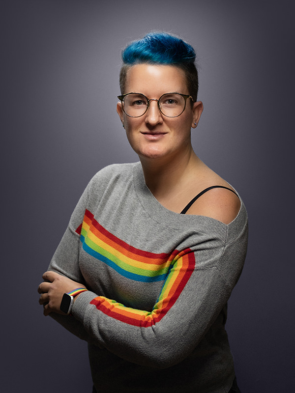 A photo of Cathie Heart, with blue hair and a grey jumper with a rainbow stripe across the front.